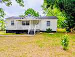 Charming single-story white house at 908 S Main Street, Louisburg, with a small front porch, black foundation, and gray roof. Surrounded by grass and trees, it features minimal landscaping plus a ramp and stairs leading to the porch.