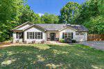 Charming single-story white house in Oxford at 4508 Alice Drive, featuring dark shutters, a front porch with two chairs, attached garage, gravel driveway, and lush green trees and grass under a clear blue sky.
