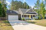 Charming single-story suburban house at 230 N Ridge Drive, Louisburg, with a front porch, attached two-car garage, manicured shrubs, and large driveway. Gray siding and white trim overlook a wooded area under a clear blue sky.