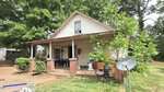 A small, single-story house at 1310 2nd Street in Henderson features a covered front porch with brick pillars and white siding. A satellite dish and chair sit in the yard, surrounded by trees and bushes. The porch holds various items and outdoor furniture.