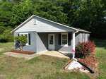 A small, single-story gray house with a white front door and trim at 4107 Old US Highway 64 E in Zebulon, surrounded by green grass, trees, a bush with red flowers, and a mulched flower bed. A black drainage pipe runs along the front walkway.
