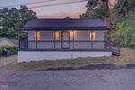 A small, single-story house at 904 Elm Street with a covered front porch, dark railings, and lit windows at dusk. The Henderson home sits slightly elevated above cracked pavement, framed by trees and bushes in the background.