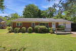 A single-story red brick house with white trim at 1315 Alpha Road in Henderson, featuring shrubs in front, a sloped roof, a carport and ramp on the right side, plus a grassy yard with trees and blue sky in the background.