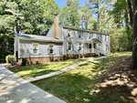 Located at 84 Wintergreen Road in Henderson, this two-story house features light gray siding, white trim, a brick foundation, and a covered front entrance. Multiple windows with shutters overlook a sloped lawn surrounded by trees.