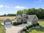Aerial view of a large brick house at 120 Barn Hill Lane, Wake Forest, with multiple gables and a three-car garage, set on a spacious lot with manicured landscaping, detached garage, backyard pool, and scenic views of trees and open fields.