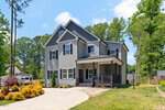 A modern two-story gray house with white trim at 127 Ottawa Drive, Louisburg, features a covered front porch, large windows, and a sloped driveway, surrounded by green trees and shrubs under a blue sky.