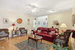 A cozy living room with a red leather sofa, wicker chairs, a wooden coffee table, floral rug, and several lamps. Light walls, wood flooring, framed art, and plants complete the inviting space.