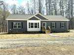 A single-story gray house with white trim, black shutters, and a small wooden front porch sits at 207 Old NC 75 S in Stem, nestled among bare trees with a dirt path leading to the entrance.