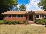 A single-story brick house at 1710 W B Street in Butner features tan shutters and a covered front porch, surrounded by trimmed bushes. The lawn is dry, with trees in the background under a partly cloudy sky.