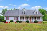 A charming one-story house at 100 Linnett Court, Youngsville, featuring gray siding, red shutters, and a spacious front porch with white railings. The yard boasts trimmed bushes, a lush lawn, mature trees, and a blue sky backdrop.