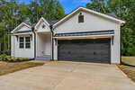 Modern single-story house with white vertical siding, black trim, and a large dark gray two-car garage. The home has a small front porch, concrete driveway, and is surrounded by trees and fresh landscaping.