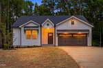 A modern single-story house at 103 Yuma Drive in Louisburg features white vertical siding, dark trim, and a double garage. Warm lights illuminate the porch and garage, with trees and a freshly mulched yard surrounding the home.