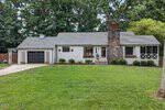 Single-story house at 1511 Oakdale Circle, Henderson, with gray siding, a dark garage door, a prominent stone chimney, and white trim. Flower boxes grace the windows, a green lawn lies in front, and tall trees rise in the background.