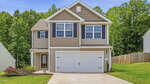 Two-story beige house with white trim at 100 Conifer Lane, Youngsville. Features a double garage, large front windows, small landscaped yard, and sits on a sloped lawn with trees and a wooden fence under a blue sky.