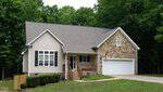 A beige house with stone accents, a covered front porch, black shutters, and a two-car garage sits on a sloped grassy lawn at 3703 Dillon Lane in Wake Forest. Trees border the yard, and an American flag hangs by the garage.