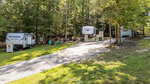 A gravel driveway in Louisburg leads past two RV campers on 171 Winnebago Loop and a small shed among tall trees. Green chairs and a fire pit sit in a grassy area beside the campers, surrounded by peaceful forest.