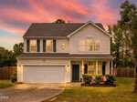 A two-story suburban house at 2085 Partridge Court, Creedmoor, with white siding, a dark roof, and a two-car garage at sunset. The front yard offers bushes, an American flag, and a tree, plus a wooden fence and trees in the background.