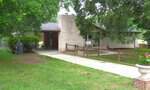 A single-story house with white brick exterior at 415 N Cheatham Street in Franklinton, featuring a large front yard, covered carport on the left, paved driveway, wooden fence near the entrance, outdoor chairs, and mature trees surrounding the property.