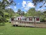A single-story house on 4298 Tommie Sneed Road in Oxford features gray siding, red shutters, and a wooden ramp leading to the front door, all set amid green grass, trees, and a blue sky with scattered clouds.