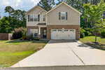Two-story suburban house at 1181 Ridgeland Drive in Creedmoor, featuring beige siding, a brick facade, and a white double garage. A driveway leads to the entrance, with a lawn bordered by plants and trees under a partly cloudy sky.