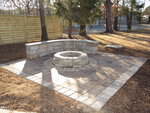 A stone fire pit with a curved stone bench sits on a square patio made of pavers, surrounded by trees and a wooden fence in a yard.