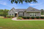 A modern gray house with white trim at 15 Ashland Court, Zebulon, features a gabled roof and large windows behind manicured bushes and a curving sidewalk, surrounded by green lawn and trees under a blue sky.