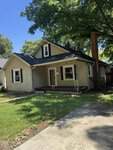 A small beige house at 212 Zollicoffer Avenue, Henderson, with a steep dark roof, covered front porch, white trim, and brick chimney sits shaded by large trees, featuring a patchy lawn and concrete driveway in front.