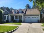 A gray suburban house with white trim at 302 Oxford Park Boulevard, Oxford, features a front porch, two dormer windows, a double garage, and is surrounded by green grass, shrubs, and trees under a clear blue sky.