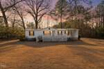 A white single-story house at 870 Jackson Royster Road in Henderson features a front porch and blue shutters, set in a spacious yard bordered by trees at sunset, with warm light glowing from its windows.
