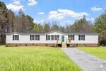 A wide, single-story white manufactured home with black shutters and a bright blue front door sits on a grassy lawn with a gravel driveway at 6631 Roundstone Lane, Oxford, surrounded by trees under a sunny sky.