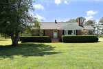 A one-story brick house with white trim and a gabled roof at 4306 Tommie Sneed Road, Oxford, features central front steps and manicured hedges on a large grassy lawn beneath a blue sky with scattered clouds.