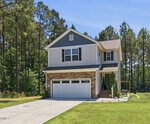 A two-story suburban house at 130 Teal Drive in Youngsville with a two-car garage, beige siding, stone accents, and black shutters, surrounded by a green lawn and tall pine trees under a clear blue sky.