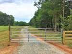 A metal farm gate blocks a gravel driveway that curves between a grassy field and dense green trees near Oxford, along 9025 & 9029 NC 96, under a cloudy sky.