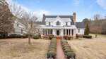 A large two-story white house at 1200 Spring Valley Lake Road, Henderson, features a covered front porch, black roof, and two brick chimneys, set on a spacious lawn with trimmed hedges and a brick walkway leading to the entrance.
