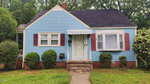 A small blue house with white trim at 214 Grace Street, Oxford, featuring maroon shutters, a white front door with an awning, brick accents at the base, lush bushes and shrubs in front, and trees in the background.