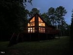 A cozy wooden cabin at 50 Tyler Court in Henderson glows warmly at dusk, its large triangular windows framed by tall trees and a grassy lawn. Stairs lead up to a spacious deck that wraps around the inviting retreat.