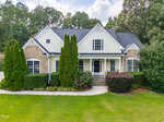 A two-story house at 509 Belmont Circle in Wake Forest features stone and yellow siding, a tall gabled roof, white trim, and a front porch. Tall shrubs and green bushes line the front, with a well-kept lawn and trees in the background.