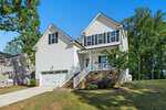 Charming two-story white house with black shutters at 102 Red Dog Drive, Louisburg, featuring an attached two-car garage, a front porch with chairs, brick steps, sloped driveway, and a grassy yard surrounded by trees.