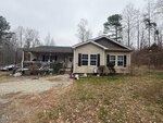 A single-story beige house with dark trim at 2700 Propus Wilkinson Road, Bullock, features two large front windows, a covered porch with plants, and a gravel driveway. Leafless trees and patchy grass surround the home under an overcast sky.