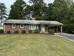 Charming single-story red brick house with white trim at 1224 Oakridge Avenue in Henderson. Features a front porch, carport on the right, green shrubs, a well-kept lawn, and tall trees in the background.