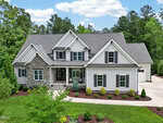 A two-story modern house at 3626 River Watch Lane, Franklinton, features gray siding, stone accents, black shutters, and a covered porch. Lush landscaping and trees surround the home, with a driveway leading to a side-entry garage.