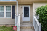 A beige house entrance with white trim, a glass door displaying the number 870, white railing, and a black shutter next to three windows. A "Warning CPI Security" sign is in the grass by the steps.