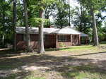 A single-story brick house with a gray roof sits among tall trees on a spacious, shaded yard at 1820 S Garnett Street Extension in Henderson. The home features a small covered front porch and several windows.