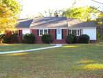 A one-story brick house at 600 18th Street, Butner, with white siding on the sides, a central front door, steps leading up, two front windows with shutters, and bushes in front. A curved walkway crosses a large grassy yard with trees in the background.