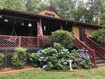 A charming wooden house at 263 Pine Trail with a covered front porch, red railings, an American flag, and blooming hydrangea bushes in the yard, surrounded by green trees and shrubs in Henderson.
