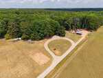 Aerial view of a rural property at 1250 Gilcrest Farm Road, Wake Forest, with a winding gravel driveway, a small garden plot, a barn, and several vehicles near woods, surrounded by open fields and dense trees under a partly cloudy sky.