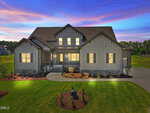 Modern farmhouse-style home at 120 Broadleaf Lane in Youngsville, featuring gray board and batten siding, stone accents, and a gabled roof illuminated at dusk. A curved driveway leads to a landscaped yard with young shrubs and a small statue out front.