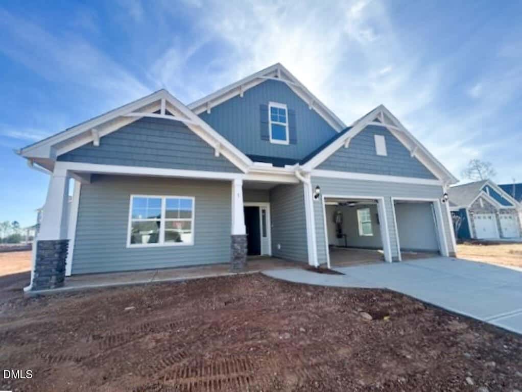 A newly built two-story gray house with white trim, stone pillars, and a covered front porch at 5 Bee Balm Court in Youngsville. Features a double garage, concrete driveway, and bare soil yard beneath a clear blue sky.