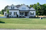 A modern two-story house at 70 Firefly Lane in Youngsville features white siding, gray accents, a dark roof, fresh landscaping, young trees, a stone walkway, and new grass, all set against a scenic wooded backdrop.