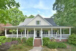 A white, two-story house at 5933 2 Pines Trail, Wake Forest, with a black roof and wraparound porch adorned with hanging plants and brick steps, sits among lush trees and bushes on a sunny day.
